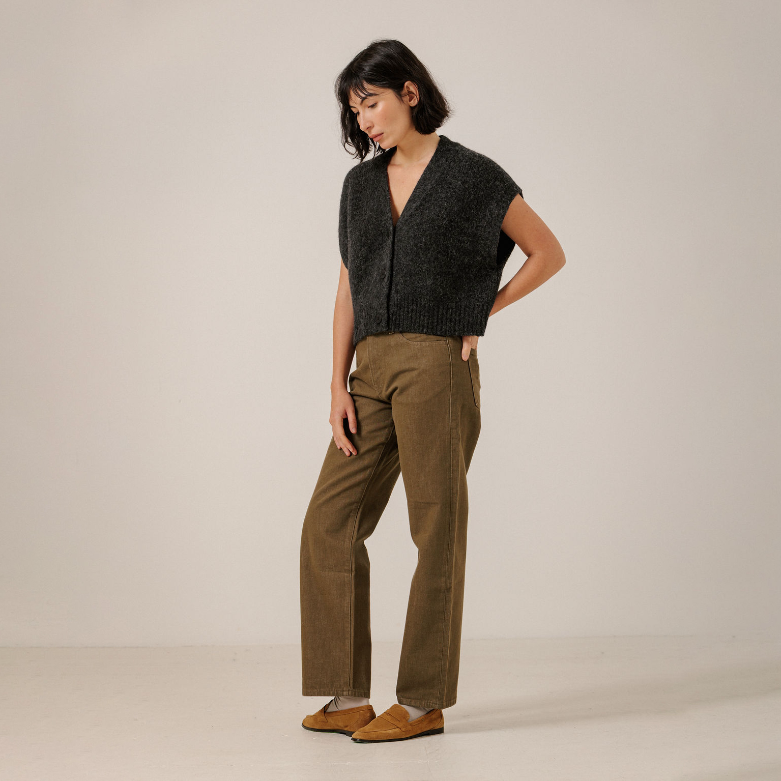 A woman stands in a studio wearing a dark charcoal gray sleeveless knit cardigan, olive brown wide-leg trousers, and tan suede loafers against a plain light beige background.