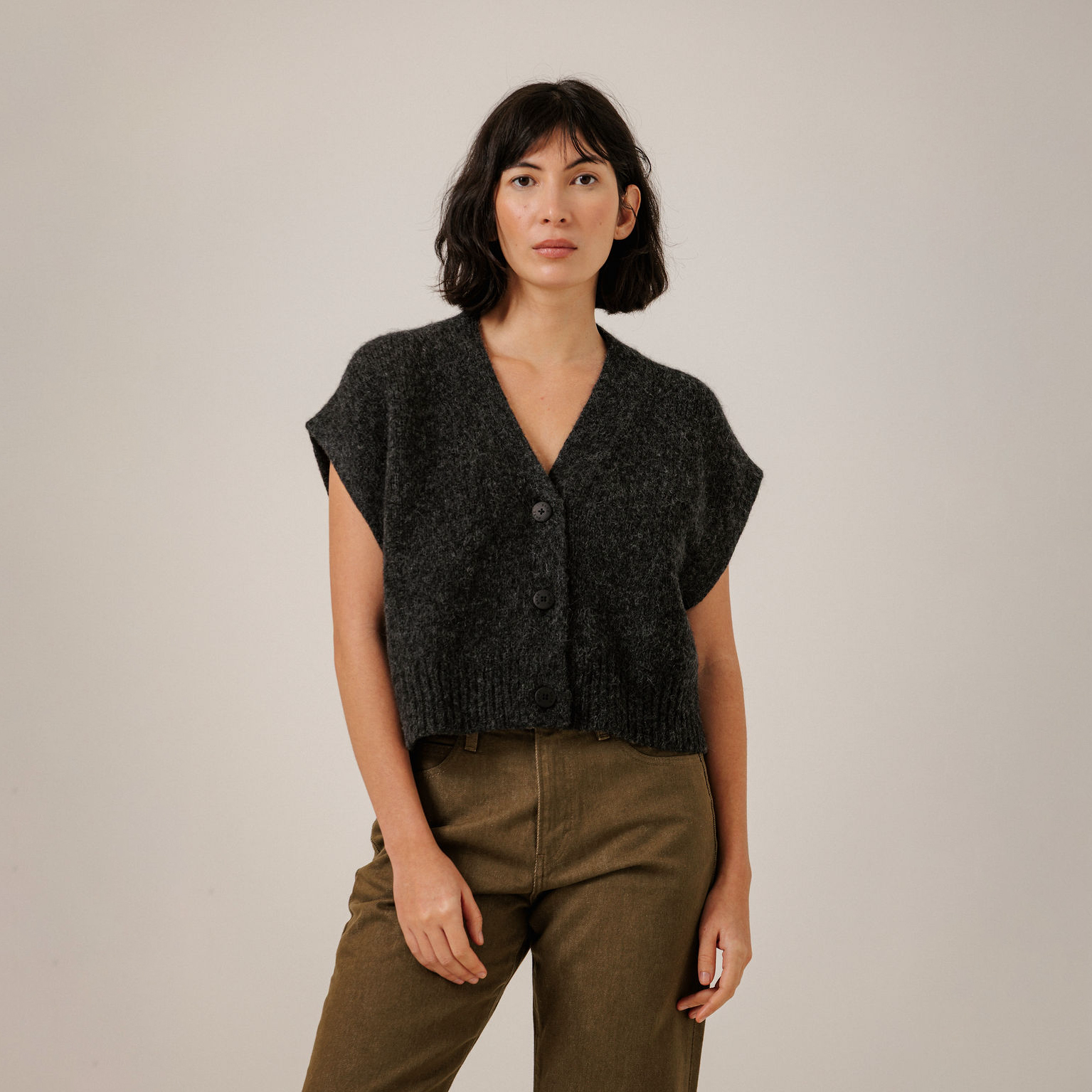 A woman modeling a dark charcoal gray knit vest with three front buttons, paired with olive green pants, against a plain light gray studio background.