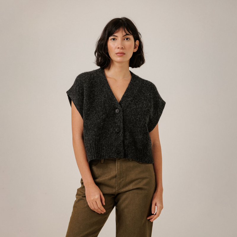 A woman modeling a dark charcoal gray knit vest with three front buttons, paired with olive green pants, against a plain light gray studio background.