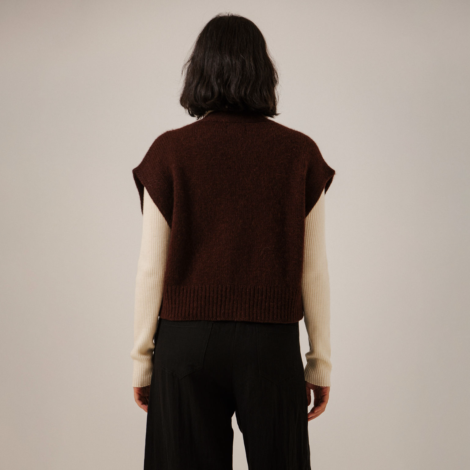 A person shown from the back wearing a dark maroon ribbed knit sweater vest over cream-colored long sleeves, paired with black pants, against a plain light beige studio background.