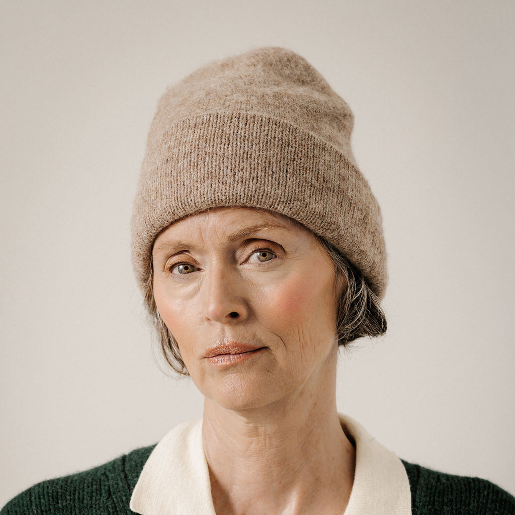 A close-up portrait of a woman wearing a beige knit beanie and a dark green sweater with a cream collar, set against a plain light background.
