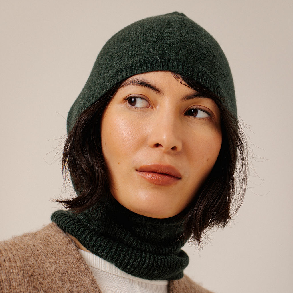 A close-up of a woman wearing a forest green knit hooded balaclava and a beige knit top against a plain light background.