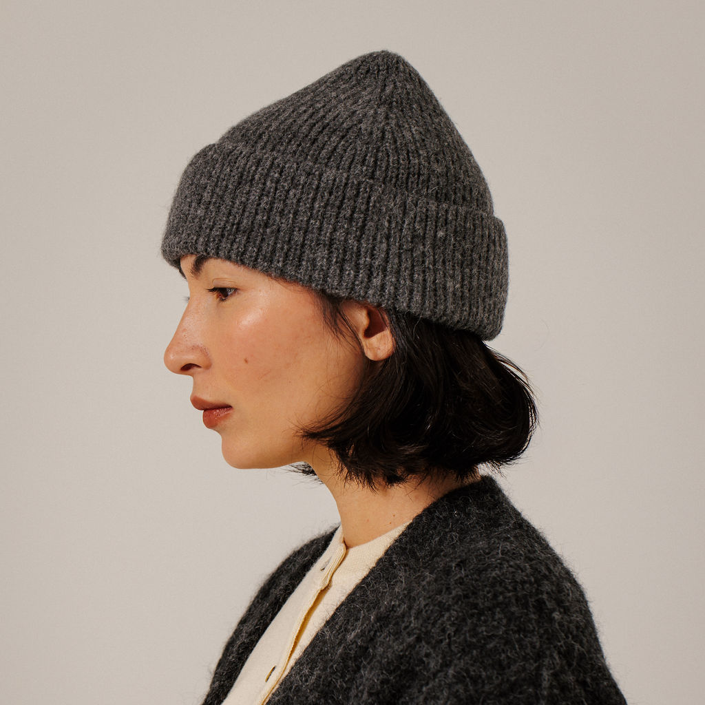 A woman in profile wearing a dark gray ribbed knit beanie and a textured dark gray cardigan, photographed against a plain light gray background.