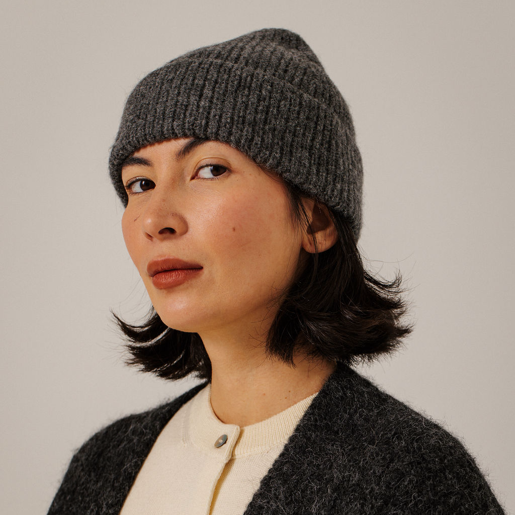 A woman wearing a dark gray ribbed knit beanie hat, shown in a close-up portrait against a plain light gray background.