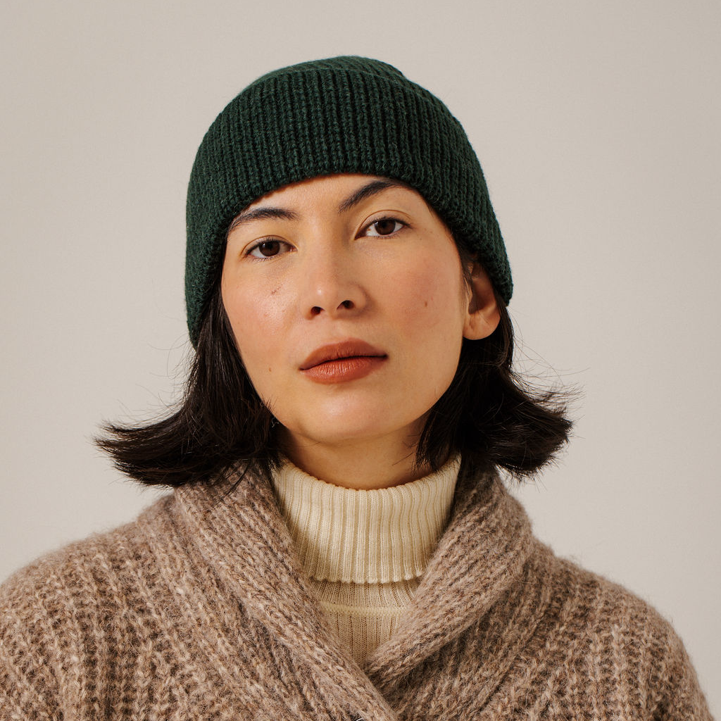 A woman wearing a dark forest green ribbed knit beanie and a chunky taupe knit sweater over a cream ribbed turtleneck, photographed against a neutral light gray background.