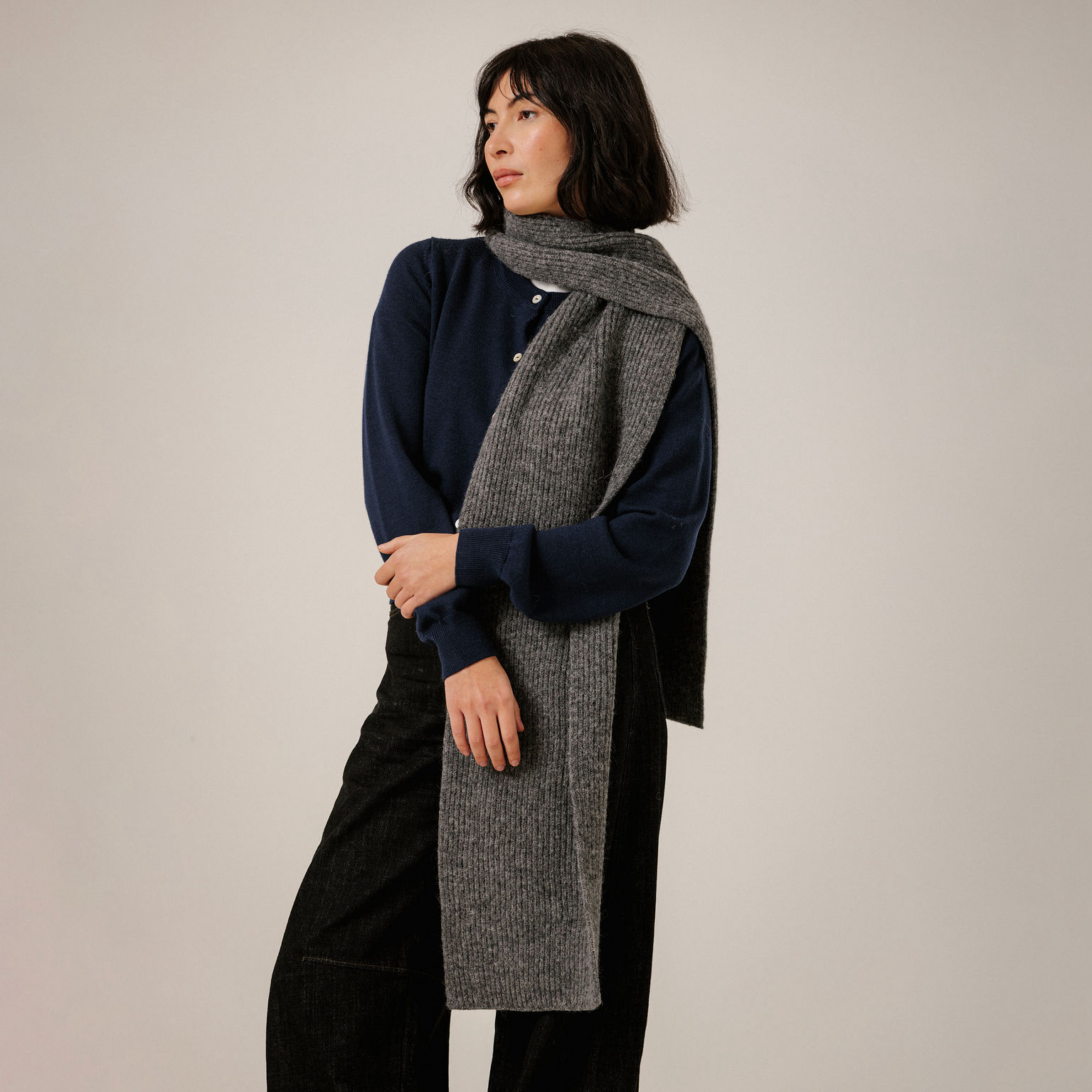 A woman modeling a long gray ribbed knit scarf wrapped around a navy blue long-sleeve top and black pants, photographed against a plain light gray studio background.