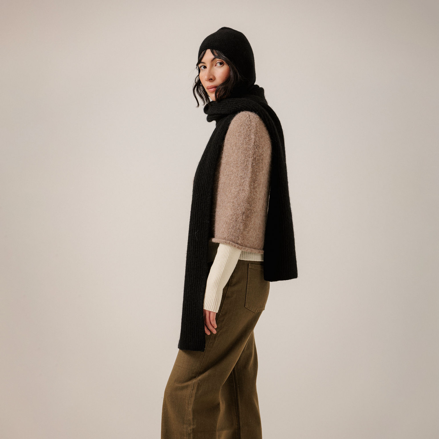 A model is shown from the side wearing a black ribbed knit scarf and matching black beanie over a beige fuzzy sweater vest, cream long-sleeve shirt, and olive green wide-leg trousers, against a plain light gray studio background.