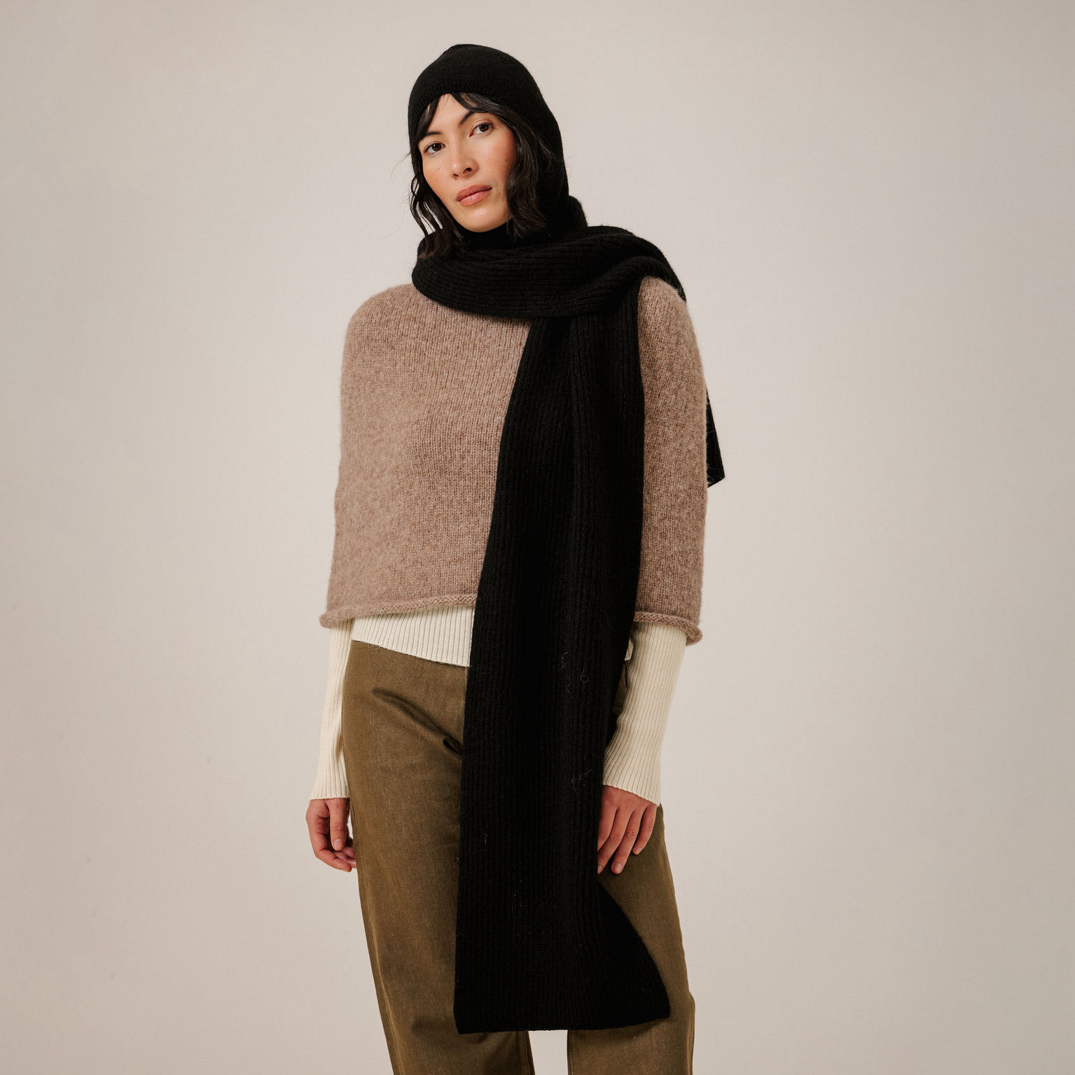A model wearing a chunky knit poncho in beige, paired with a long black ribbed scarf, a black beanie, and olive green trousers, set against a plain light gray studio background.