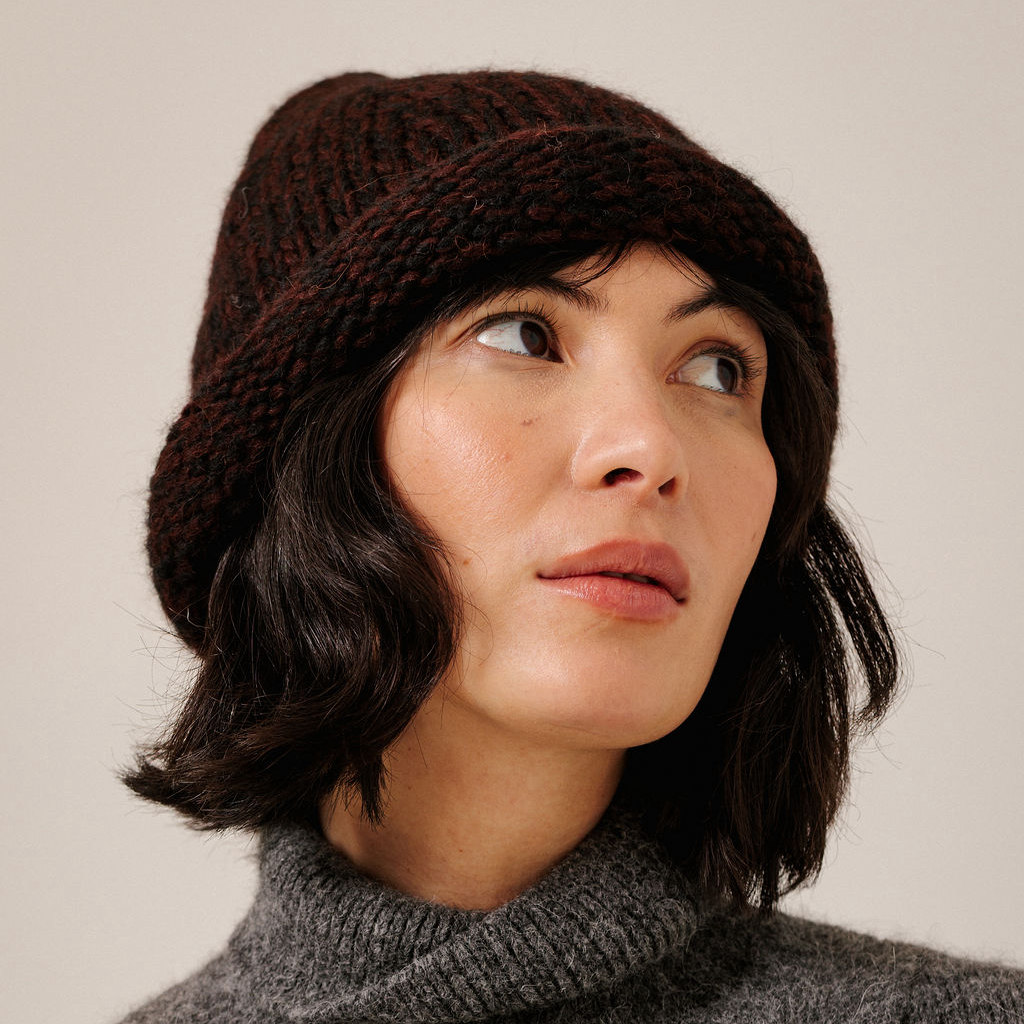 A close-up of a woman wearing a dark maroon chunky knit beanie hat and a gray turtleneck sweater, looking off to the side against a plain light beige background.