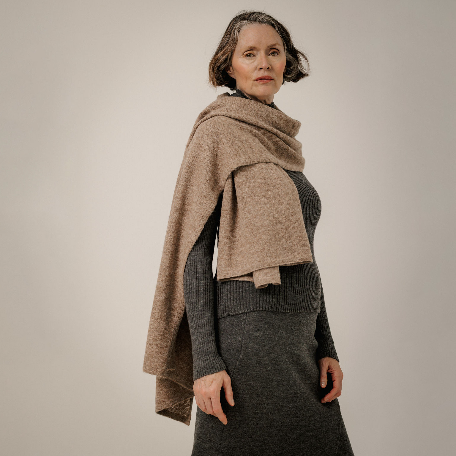 A woman wearing a taupe knit wrap shawl draped over her shoulders and a dark charcoal ribbed knit dress, photographed against a neutral light beige studio background.
