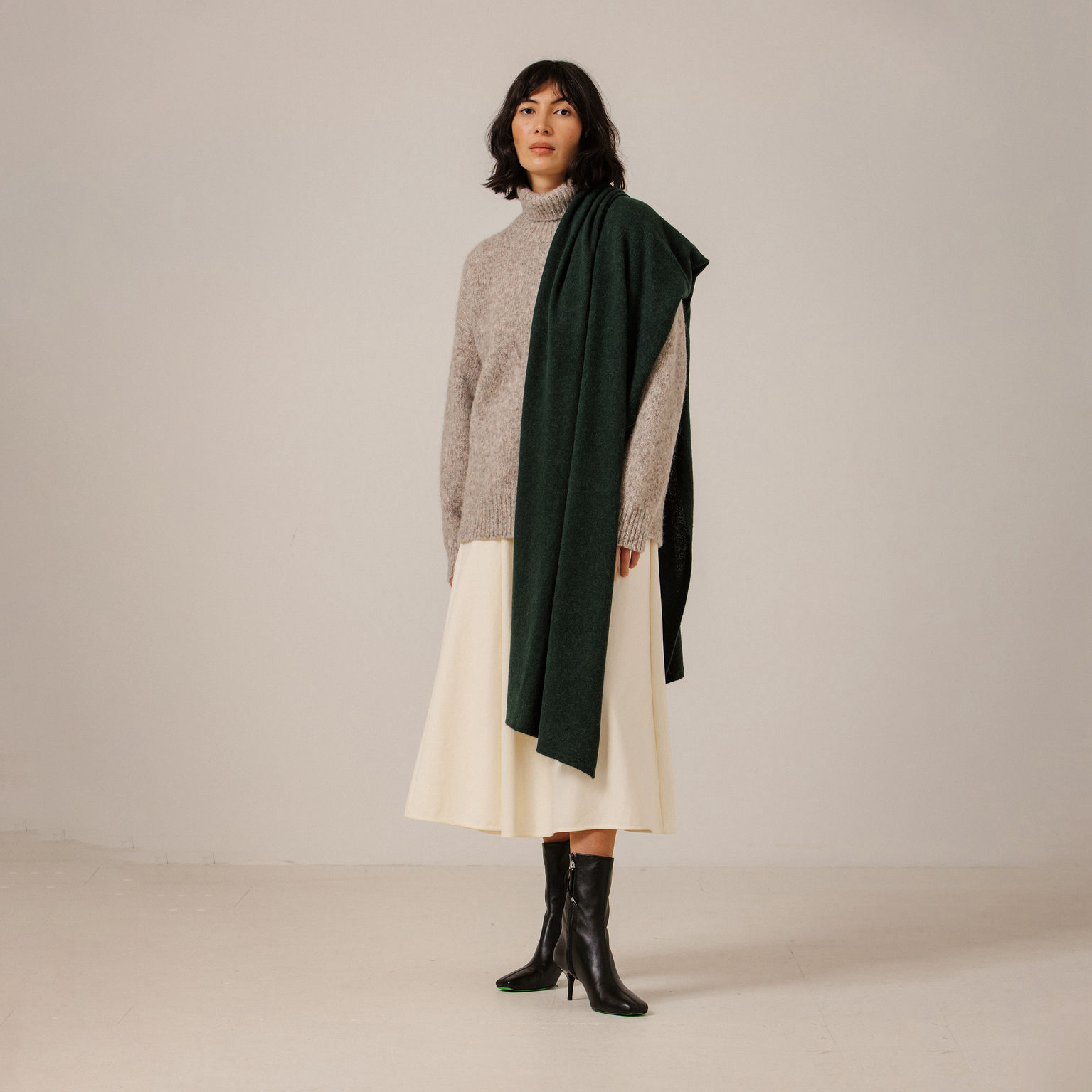 A model stands in a studio wearing a light gray knit turtleneck sweater, a cream midi skirt, black ankle boots, and a dark forest green scarf draped over her shoulders.