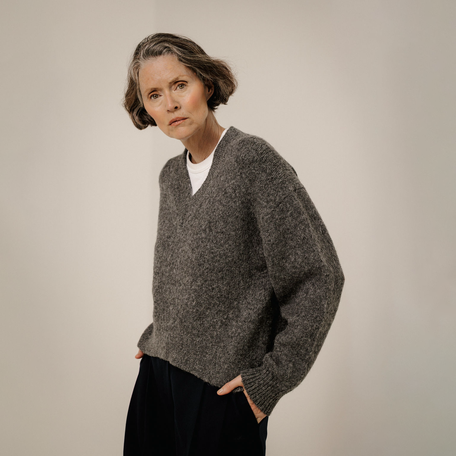 A woman wearing a dark gray heathered knit v-neck sweater over a white undershirt and dark trousers, standing against a plain light beige studio background.