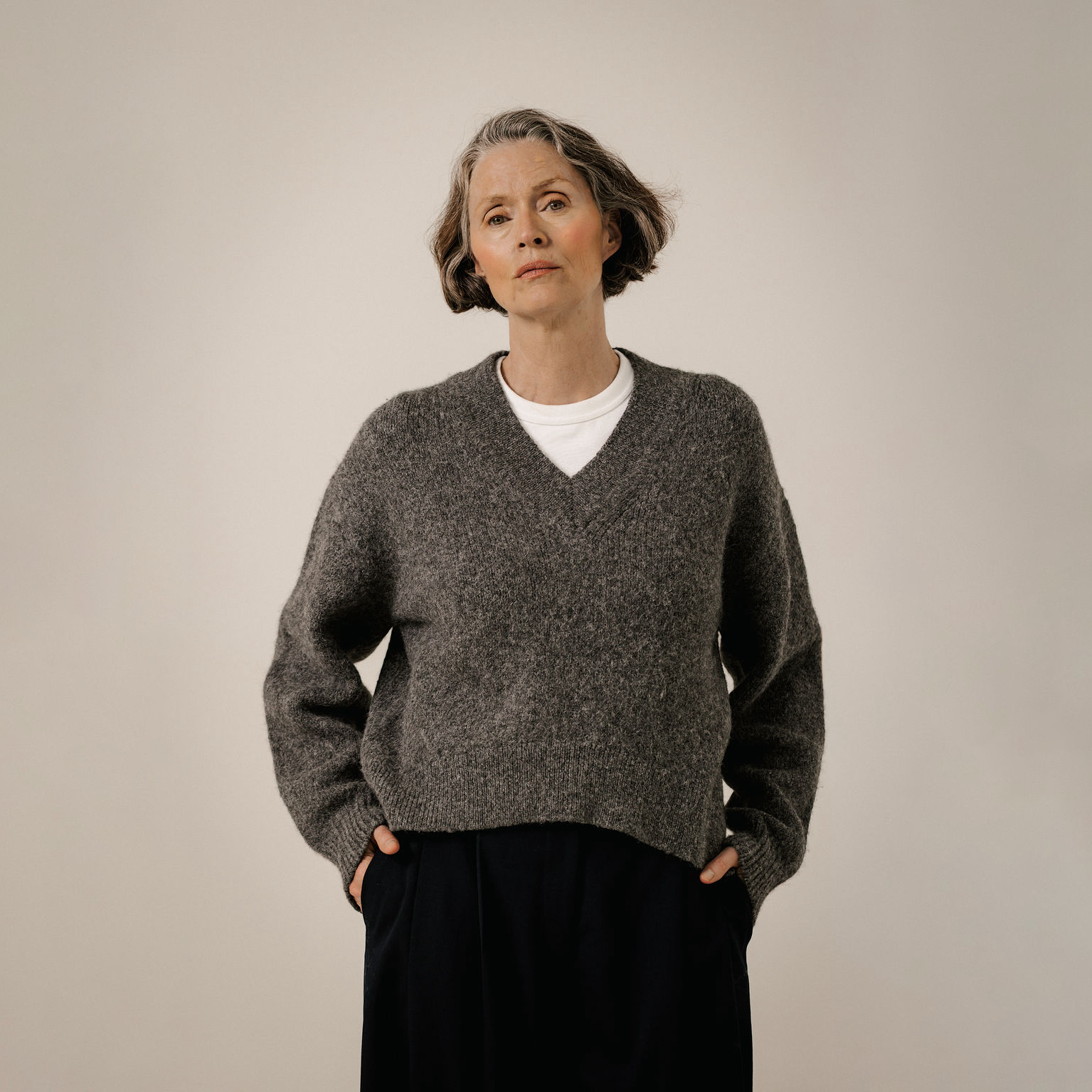 An older woman with short gray hair stands against a plain light beige studio background, wearing a dark heather gray V-neck knit sweater over a white undershirt and dark navy pants.