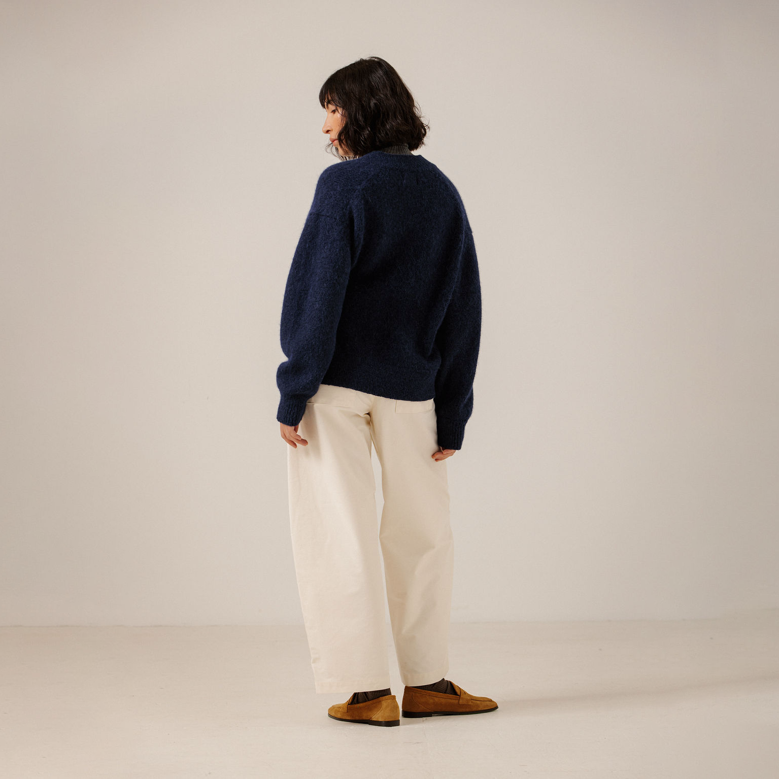 A model is shown from the back wearing a dark navy blue knit sweater, off-white wide-leg trousers, and brown suede loafers, standing against a plain light beige studio background.