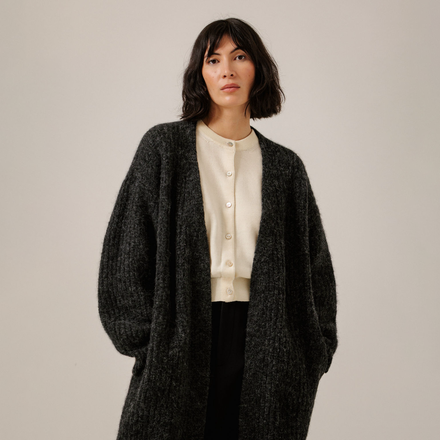 A woman wearing a dark charcoal gray textured open-front cardigan over a cream-colored button-up top and black pants, standing against a plain light beige studio background.