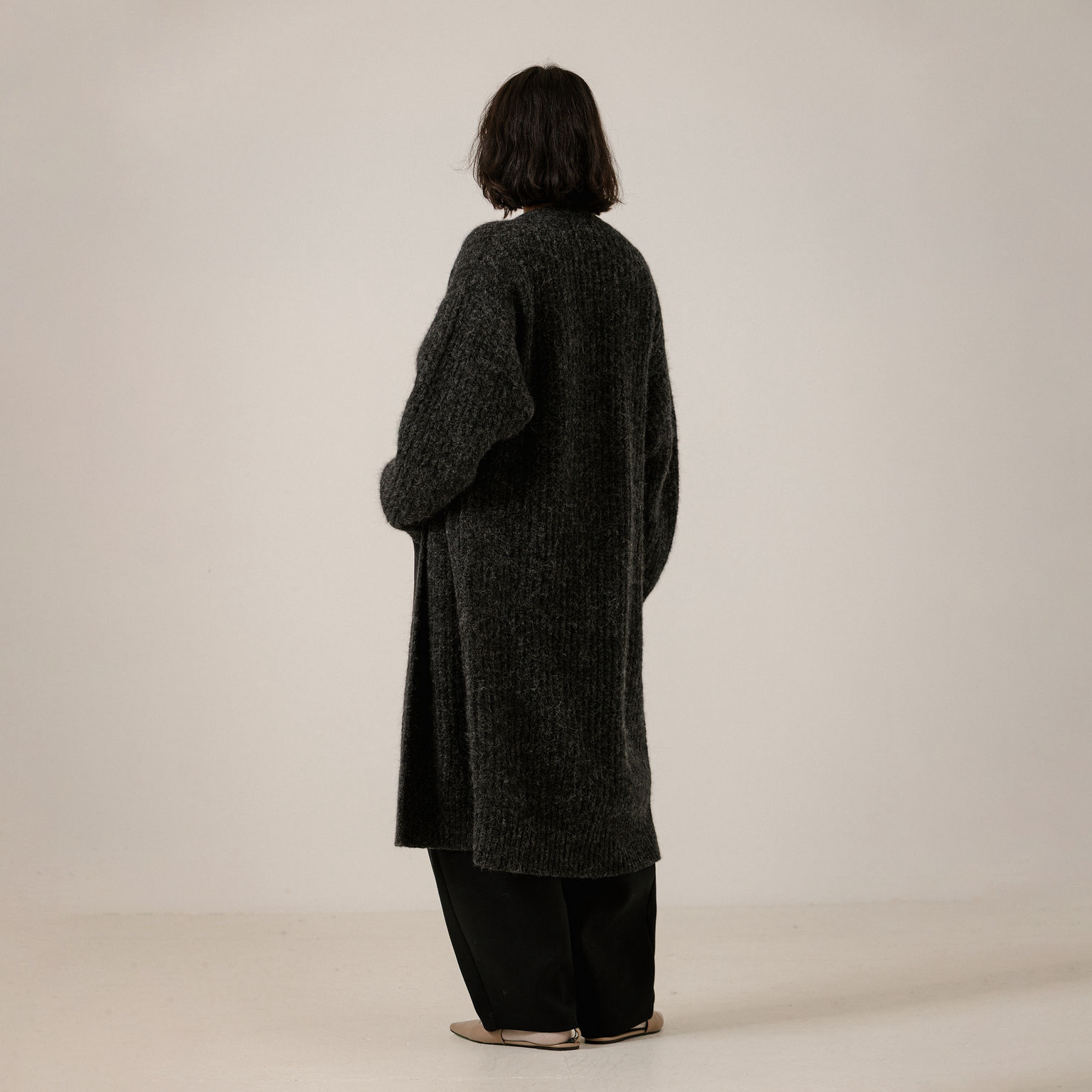 A person shown from the back wearing a long, loose, dark charcoal gray knit cardigan over black wide-leg pants, standing against a plain off-white studio background.