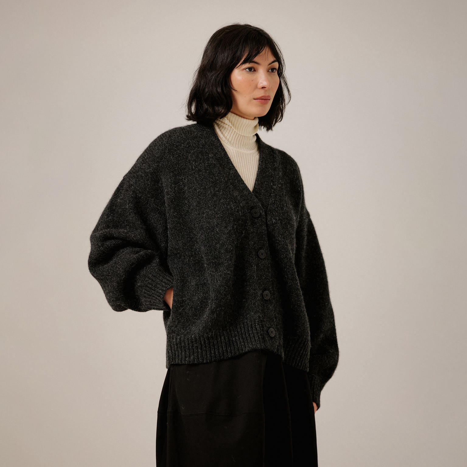 A woman wearing a dark charcoal gray knit cardigan over a cream turtleneck and black skirt, standing against a plain light gray studio background.