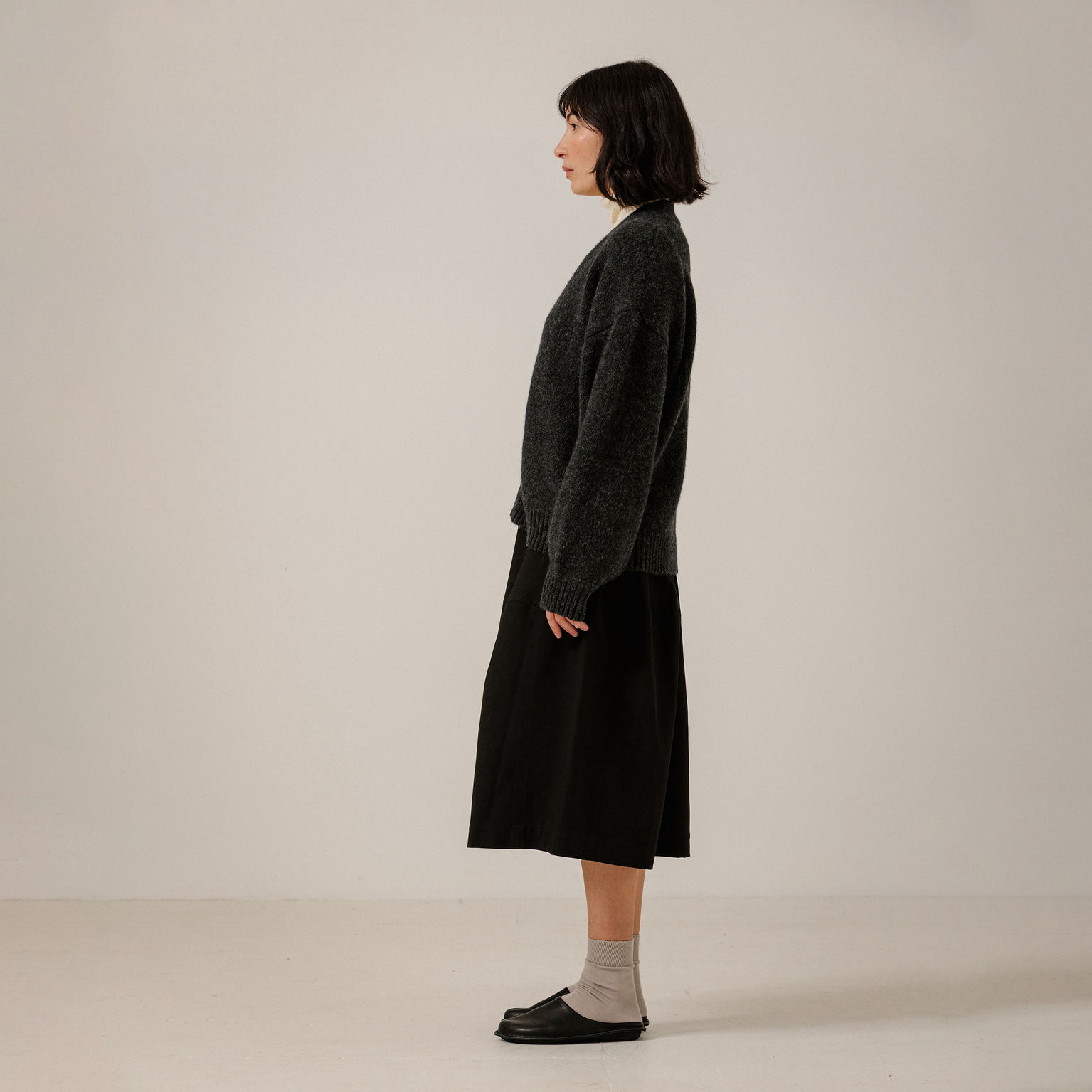 A woman stands in profile wearing a dark charcoal fuzzy knit cardigan over a black midi skirt, paired with light gray socks and black loafers, against a plain off-white studio background.