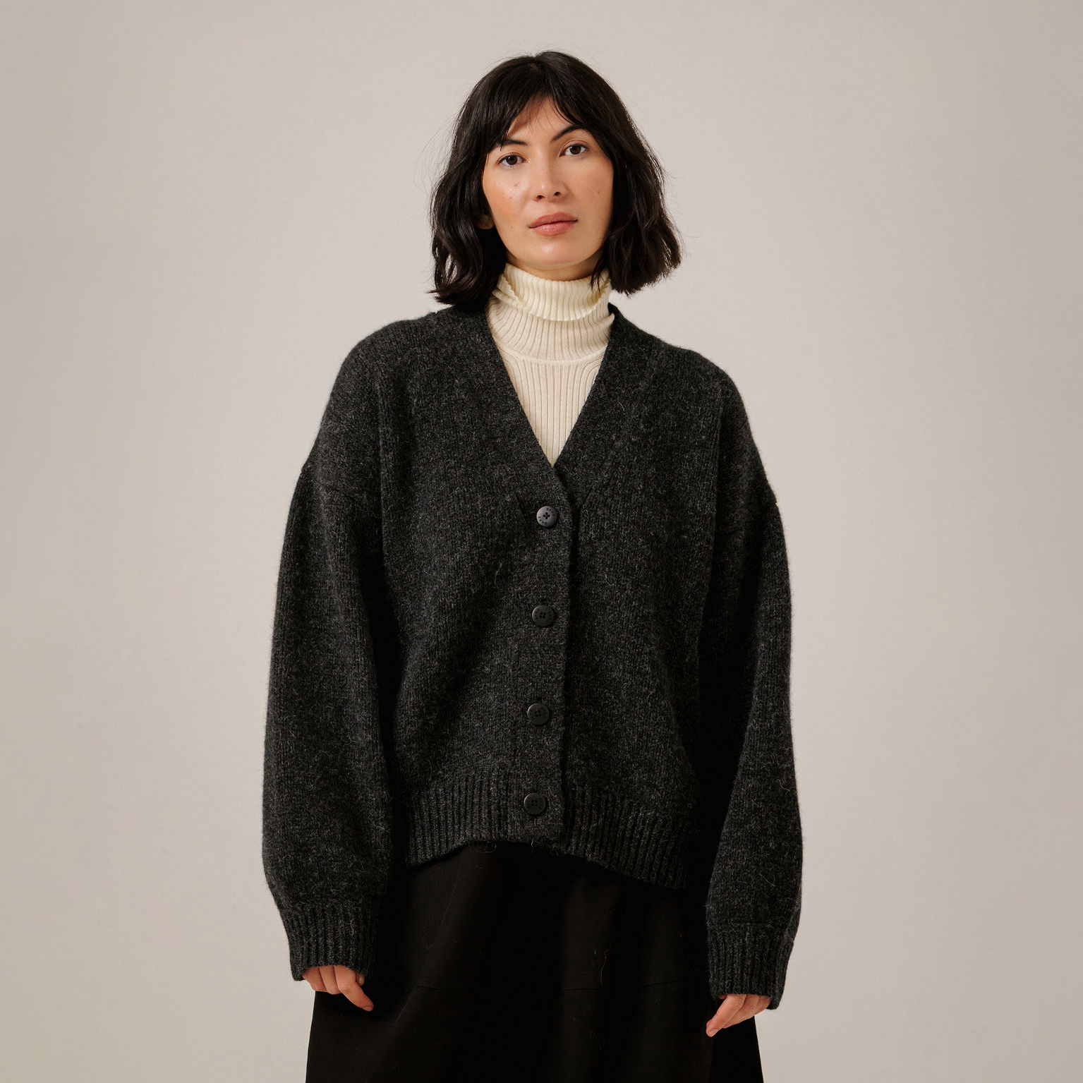 A woman wearing a dark charcoal gray knit button-up cardigan over a cream ribbed turtleneck and black bottoms, standing against a plain light gray studio background.