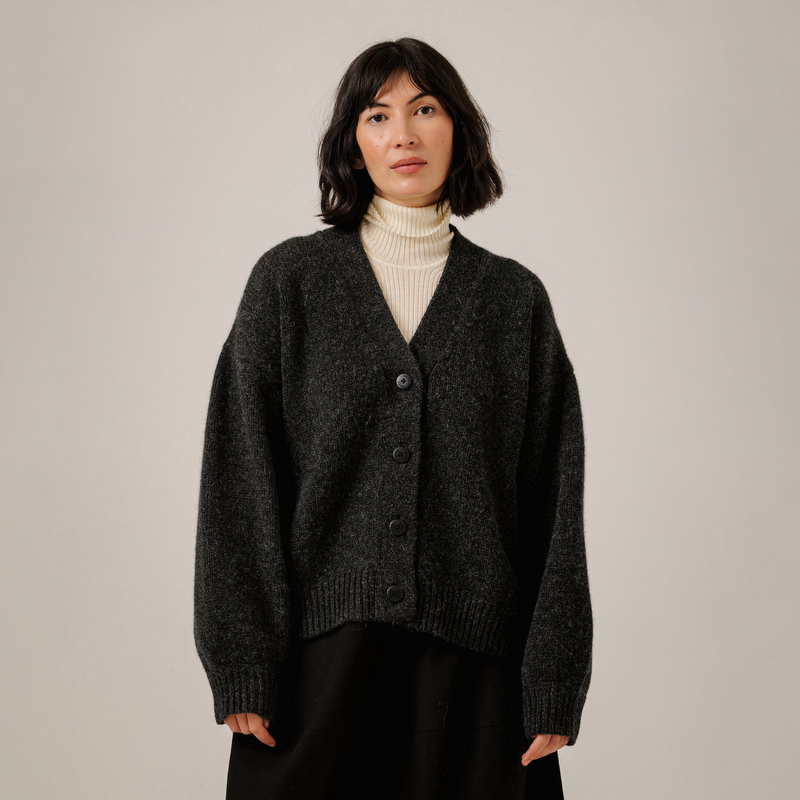 A woman wearing a dark charcoal gray knit button-up cardigan over a cream ribbed turtleneck and black bottoms, standing against a plain light gray studio background.