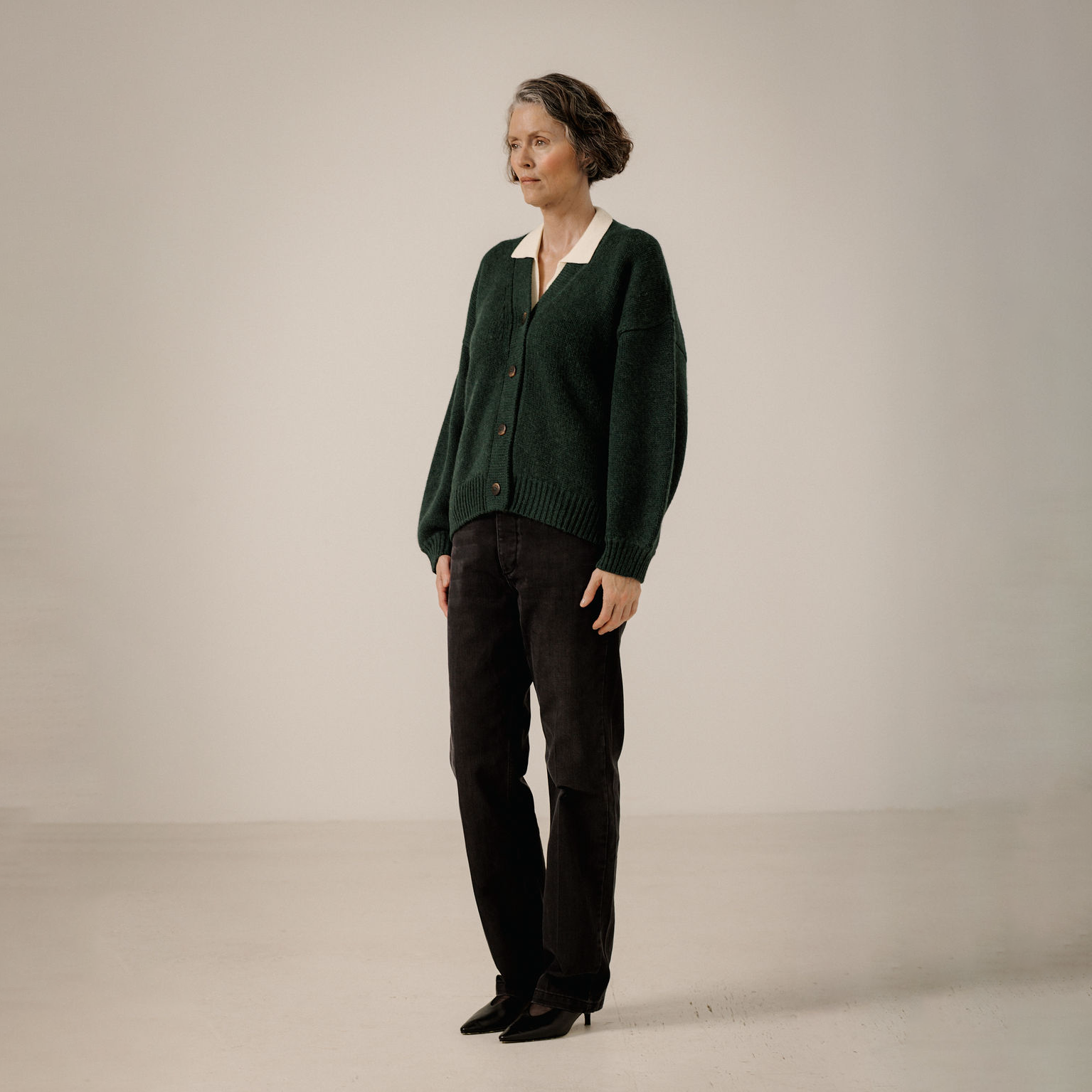 A woman stands in a studio wearing a dark green knit cardigan over a cream collared shirt, paired with black trousers and black pointed shoes, against a plain light beige background.