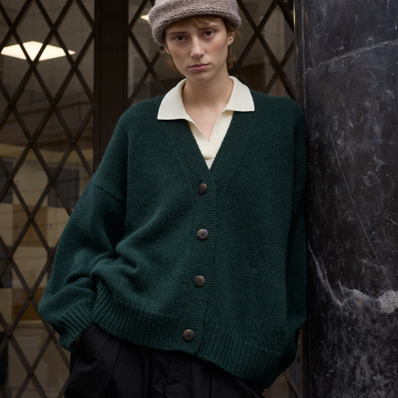 A model wearing an oversized dark green knit cardigan with brown buttons, layered over a cream collared shirt, black trousers, and a beige knit beanie, standing next to a dark marble column.