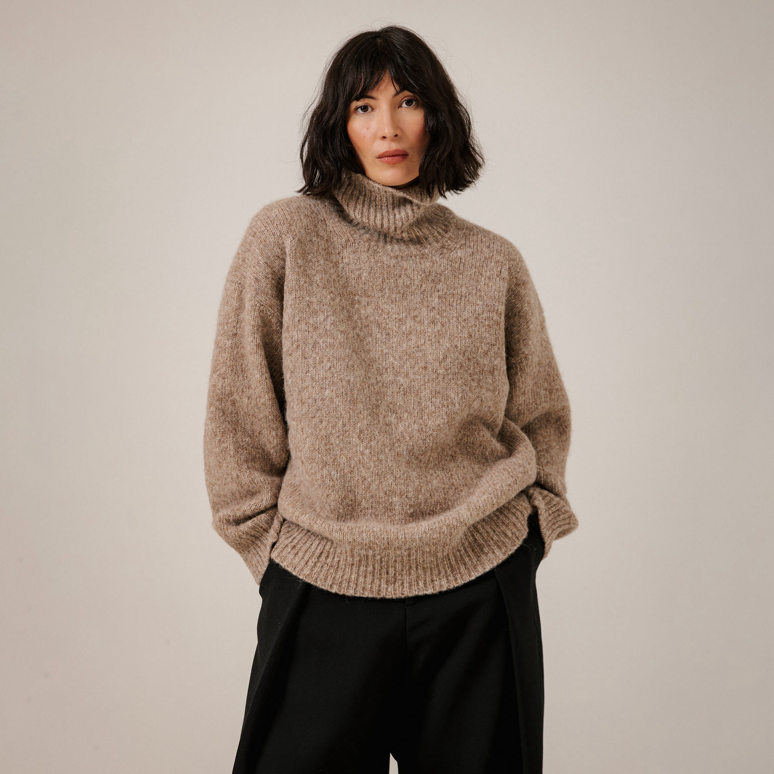 A woman wearing a chunky knit turtleneck sweater in a warm taupe-brown color, paired with black trousers, standing against a plain light beige studio background.