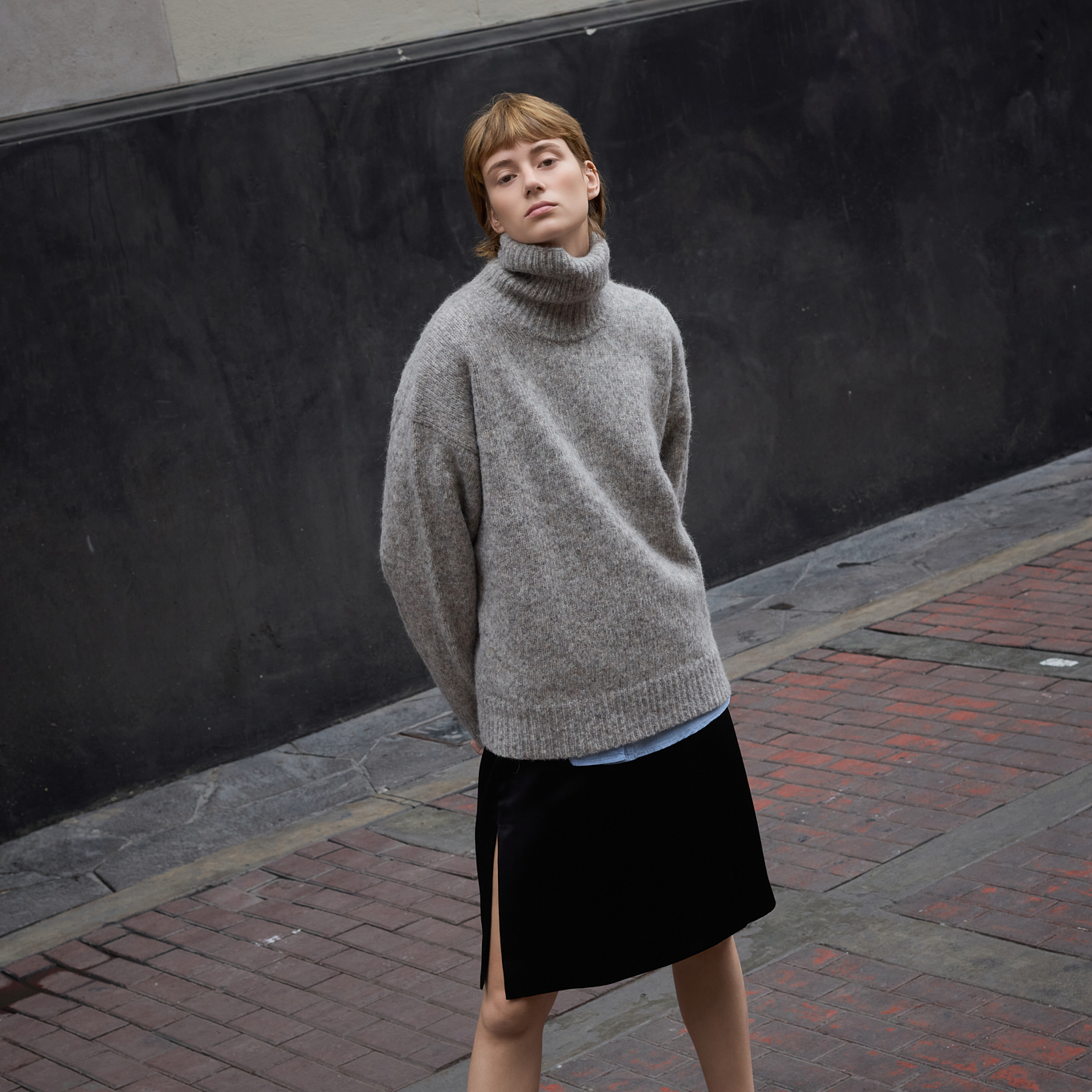 A woman wearing a chunky light gray turtleneck sweater and a black skirt, standing on a brick sidewalk against a dark wall.