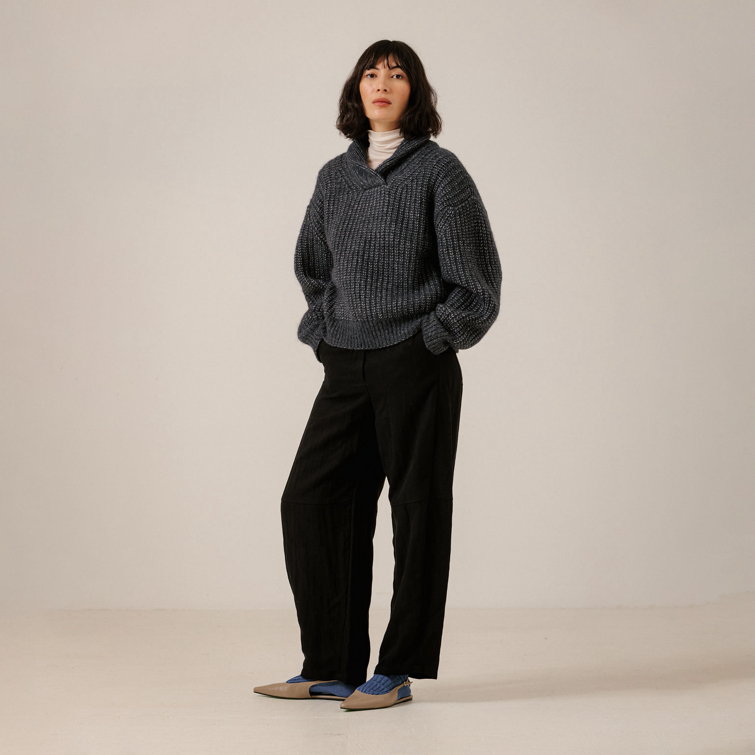 A model stands against a plain light beige studio background wearing a dark navy ribbed knit sweater over a white turtleneck, black wide-leg trousers, and blue-and-beige loafers.