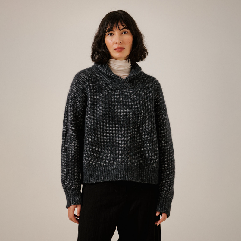 A woman wearing a dark navy textured knit sweater with a wide cowl neck, layered over a light beige turtleneck and black pants, standing against a plain light gray studio background.