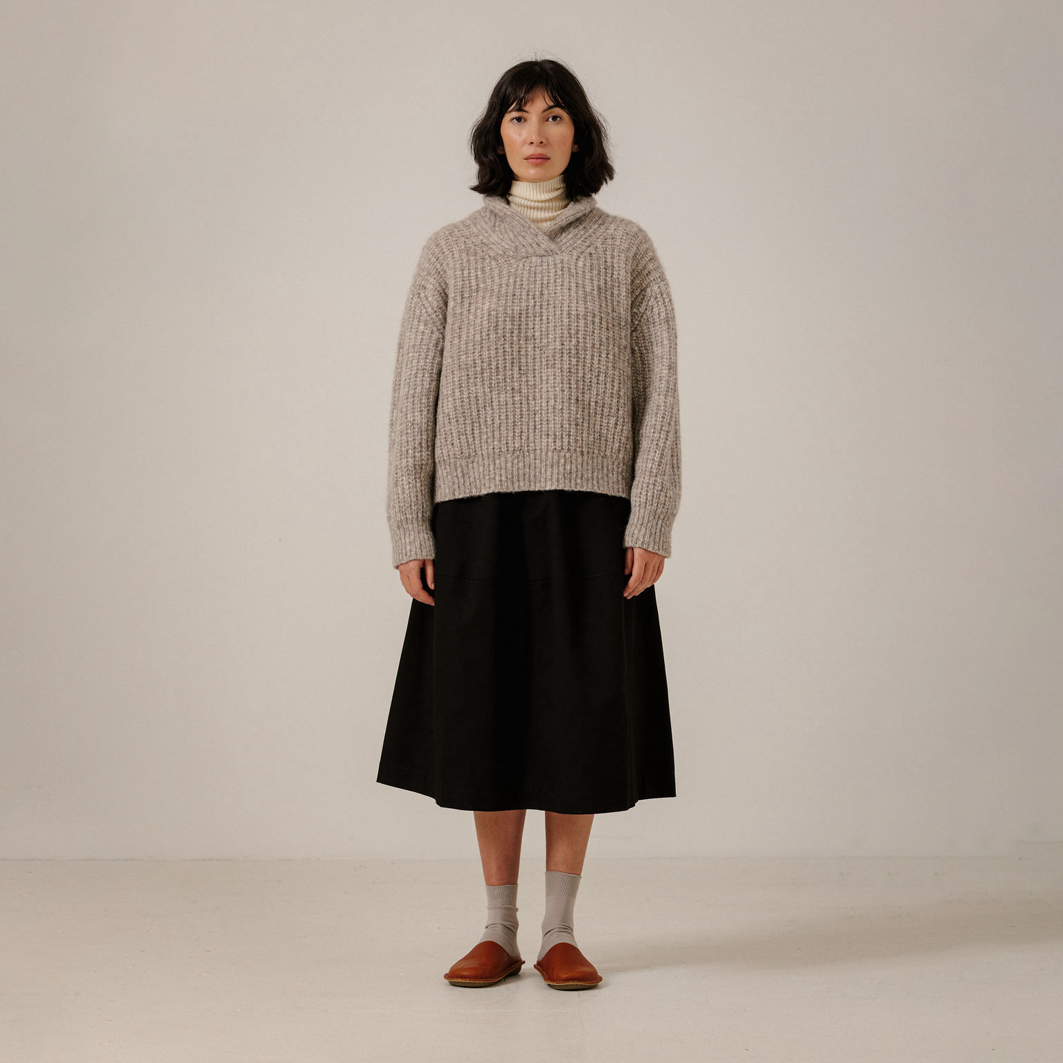 A model stands against a plain light beige studio background wearing a chunky cable-knit shawl collar sweater in a light taupe-beige color, paired with a black midi skirt, light gray socks, and brown leather loafers.