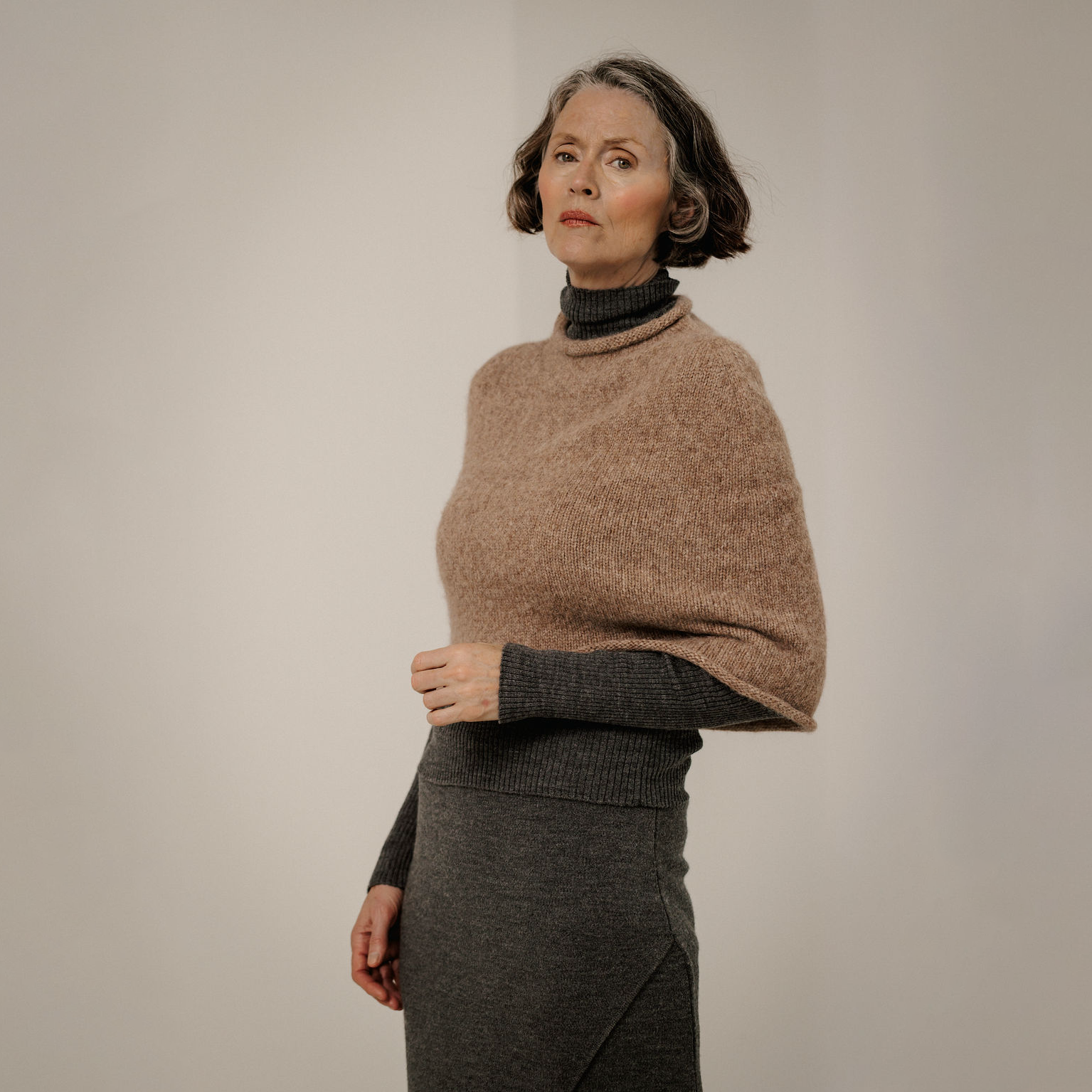A woman wearing a taupe knit poncho over a dark gray turtleneck sweater and a dark gray skirt, standing against a plain light gray studio background.