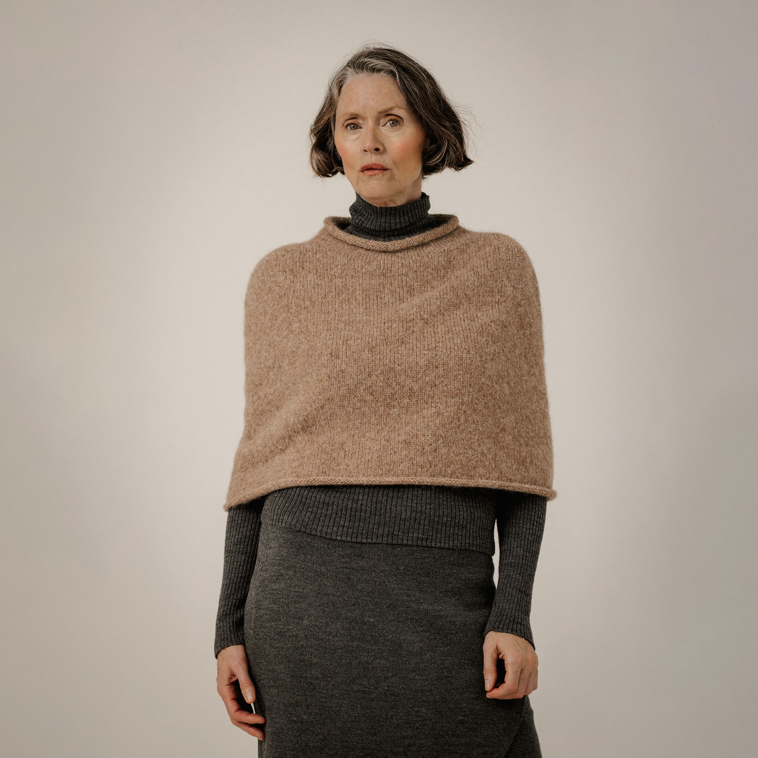 A woman wearing a chunky knit beige poncho over a dark gray turtleneck sweater and matching dark gray skirt, photographed against a neutral light gray studio background.