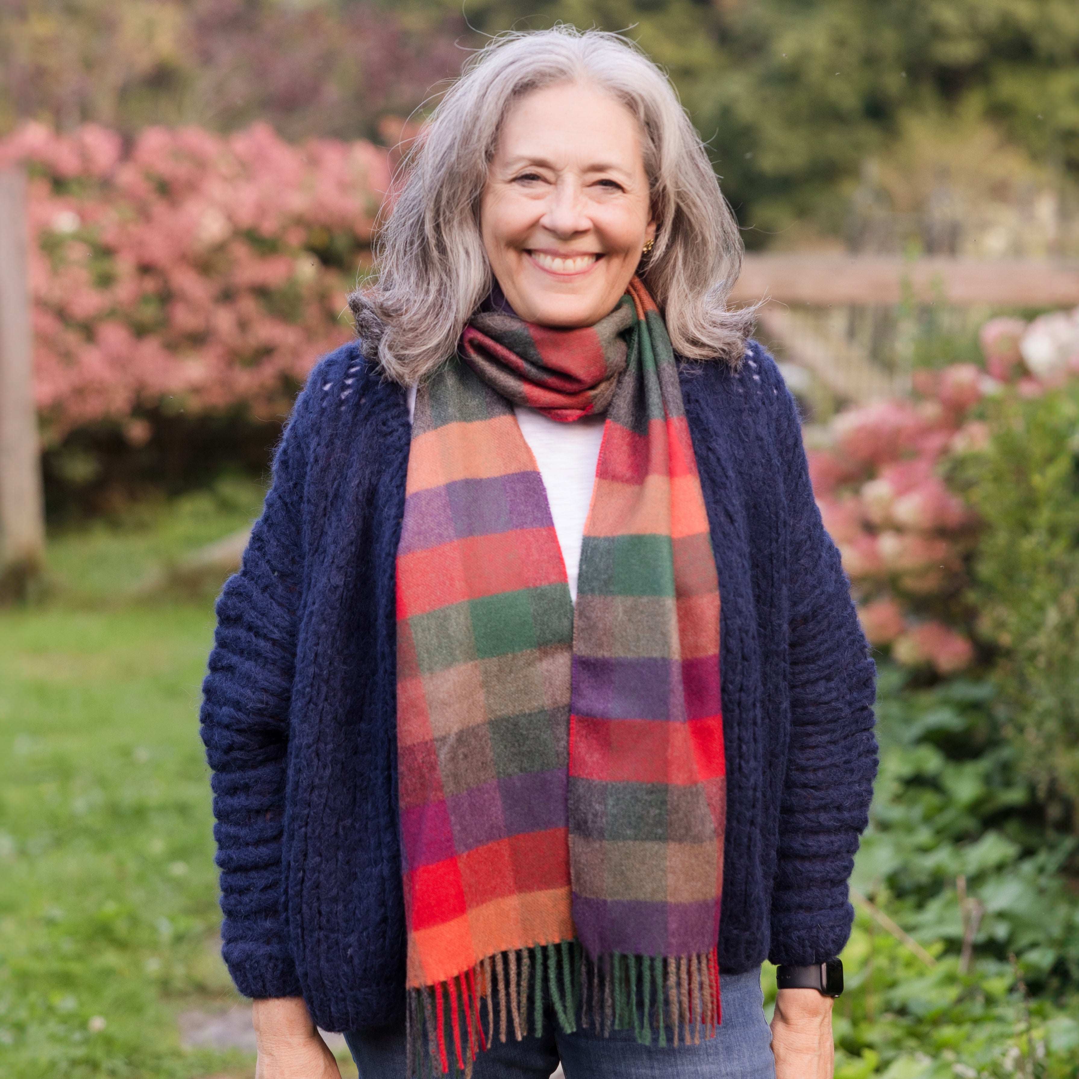 An older woman with gray hair smiling, wearing a multicolored plaid scarf with tassels, a navy blue cardigan, and a white top, standing outdoors with green and pink foliage in the background.