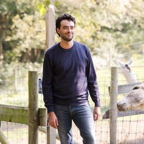 Men's Classic Crewneck 100% Baby Alpaca Sweater. A man wearing a blue baby alpaca crewneck sweater stands in front of a farm fence.