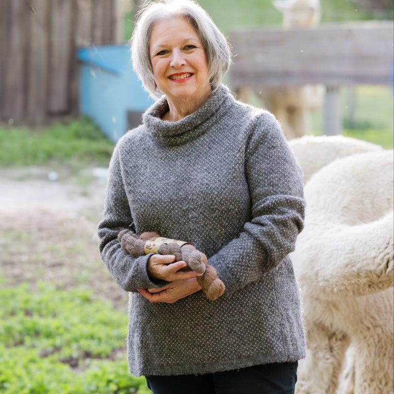 A woman standing outdoors wearing a gray mock turtleneck alpaca sweater.