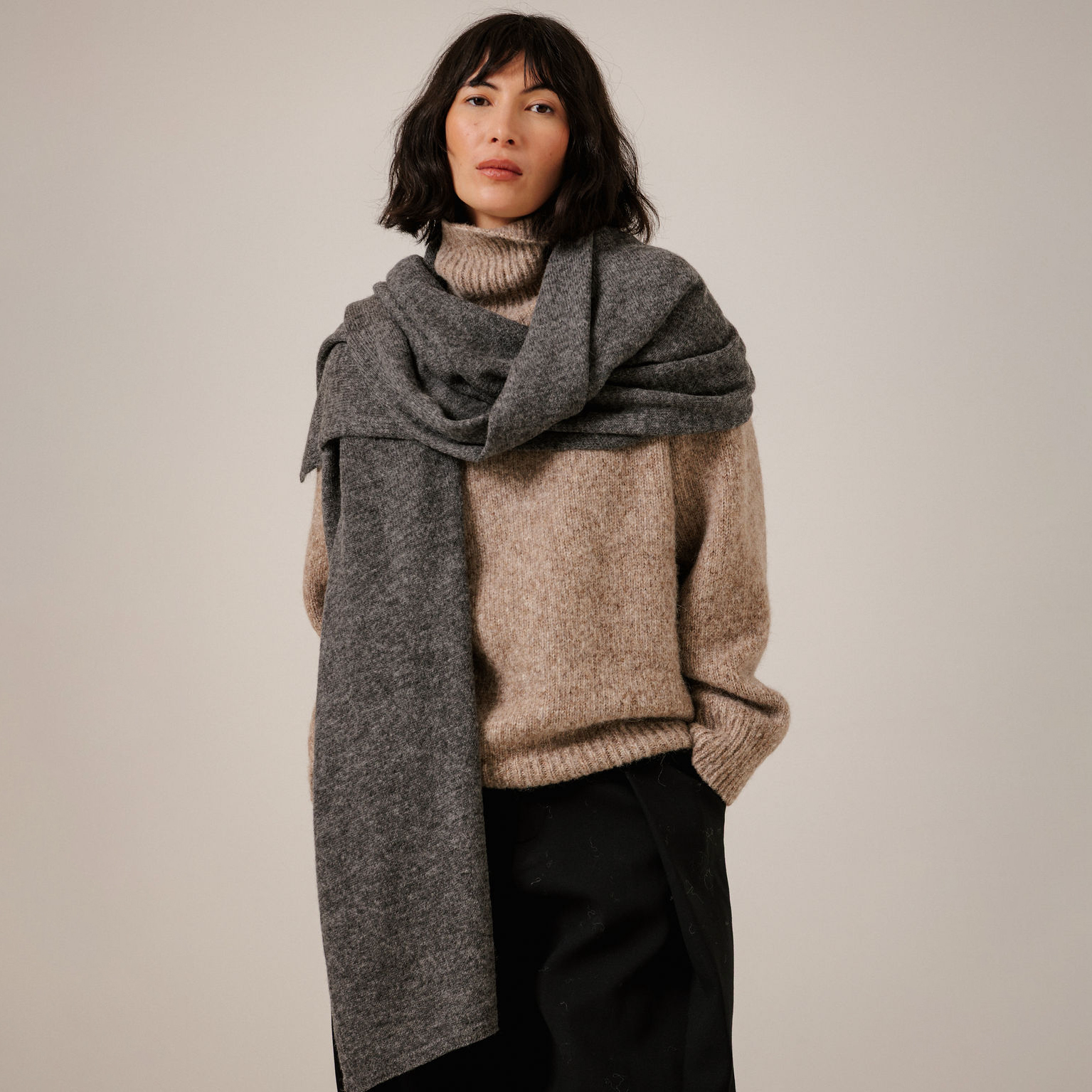A woman wearing a thick, heathered dark gray scarf wrapped around her shoulders, a beige knit sweater, and black pants, photographed against a plain light gray studio background.