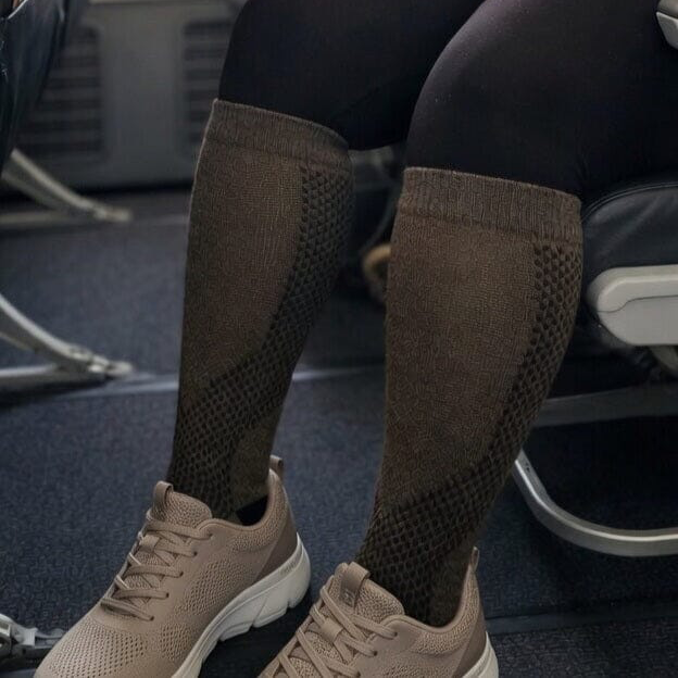 A person seated on a chair wearing knee-high knit socks in a dark brown color with textured knit panels, paired with beige athletic shoes.