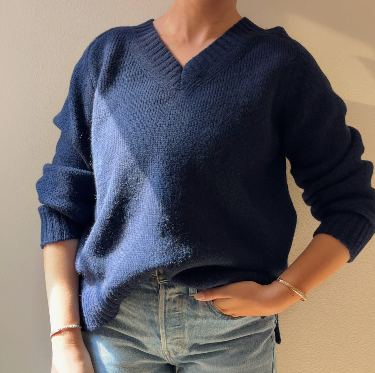 A person wearing a navy blue V-neck knit sweater with light blue jeans, standing against a plain beige wall.