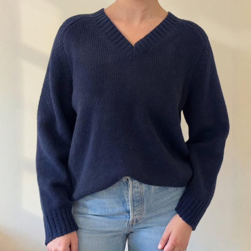 A person wearing a navy blue V-neck knit sweater with light blue jeans, standing against a plain beige wall.