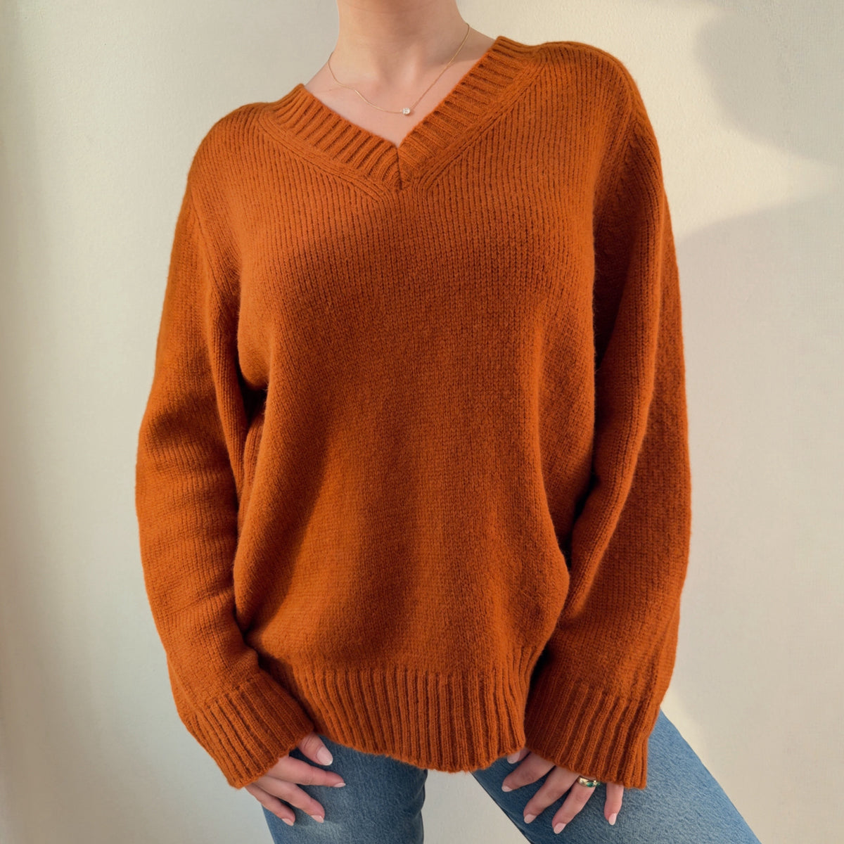 A person wearing an oversized ribbed knit V-neck sweater in a warm burnt orange color, paired with blue jeans, standing against a plain light beige wall.