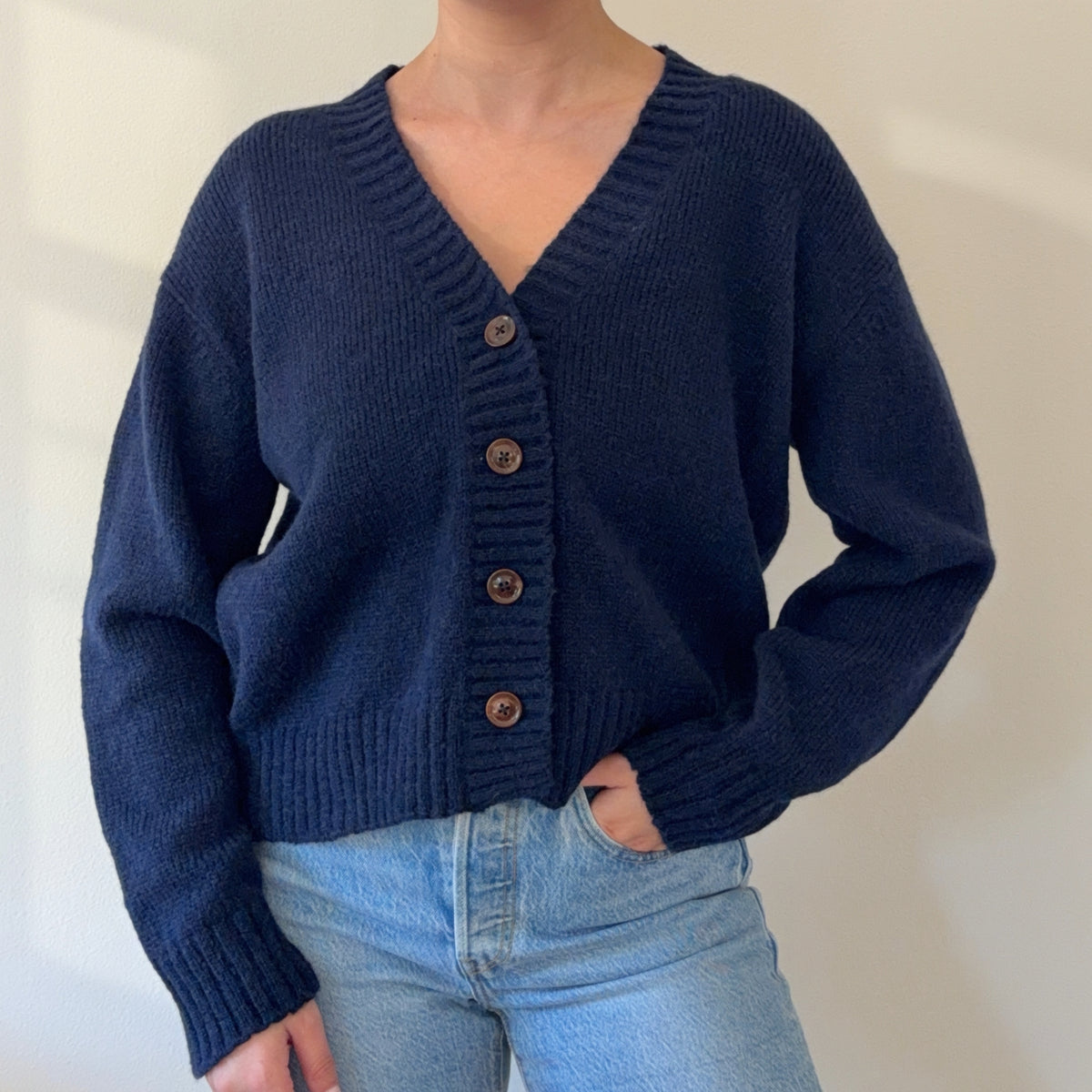 A person wearing a navy blue knit cardigan with brown buttons, paired with light blue jeans, photographed against a plain light wall.