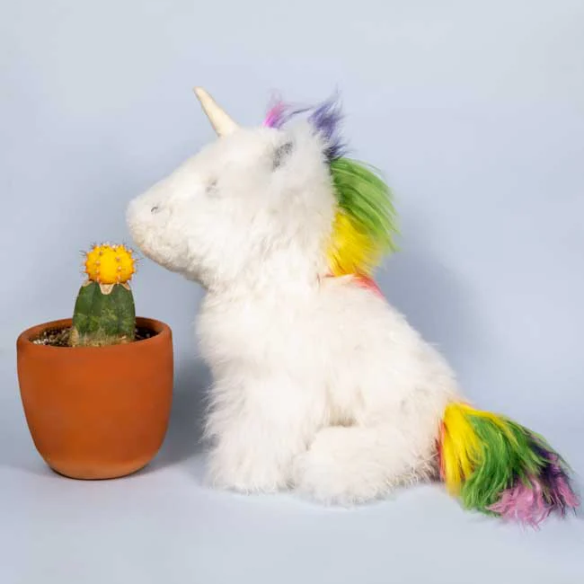 Stuffed unicorn with a rainbow mane and tail next to a potted cactus on a light gray background