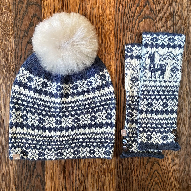A navy and white fair isle pattern alpaca wool hat with a detachable fur pom pom and matching fingerless gloves laid flat.