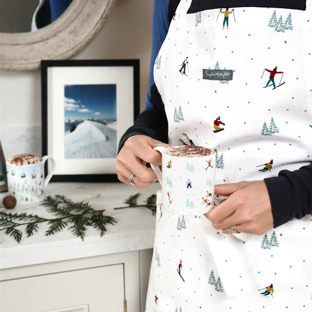 Person wearing a white apron with skiing designs, holding a mug with a hot chocolate drink.