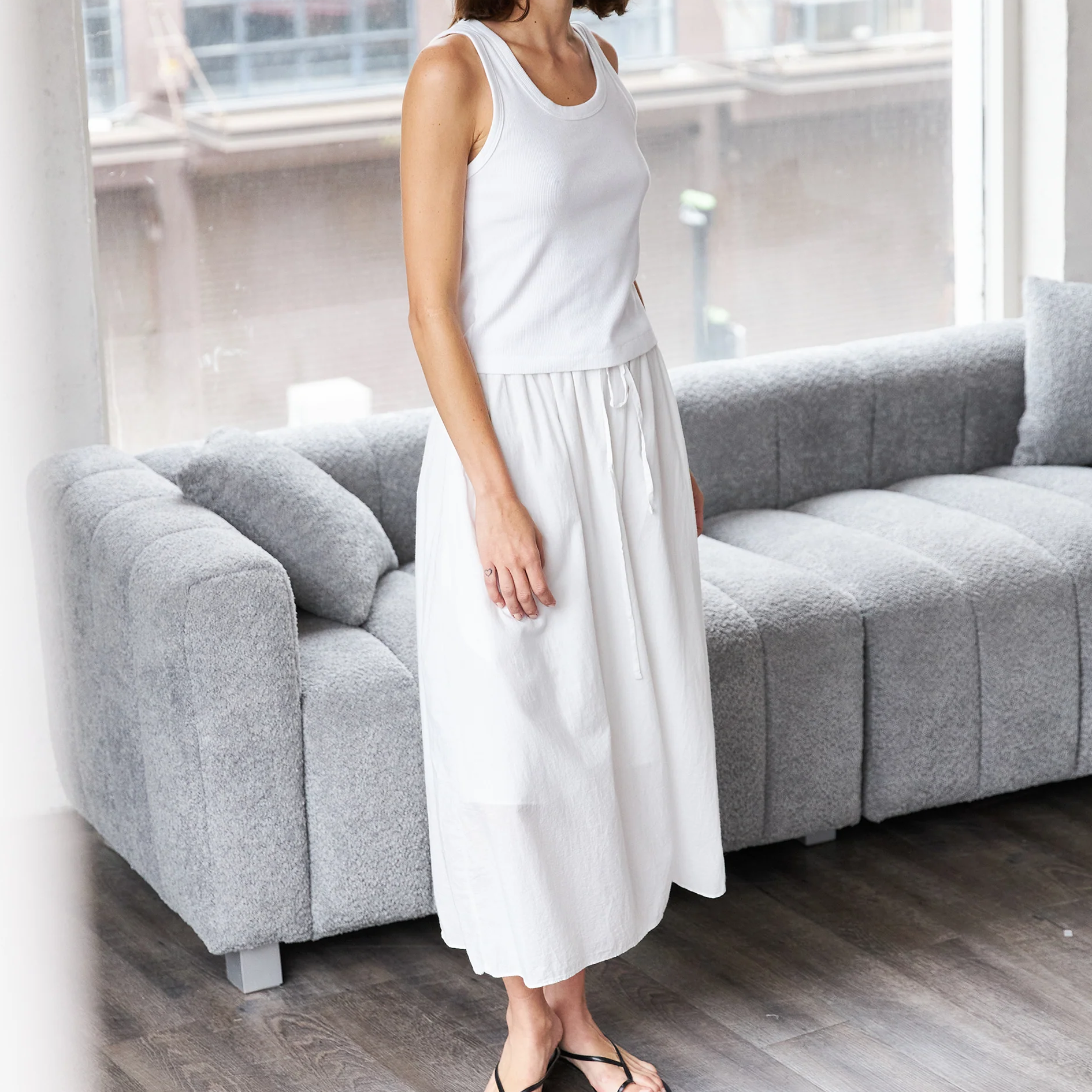 Woman in a white sleeveless top and skirt standing in a room with a gray sofa.
