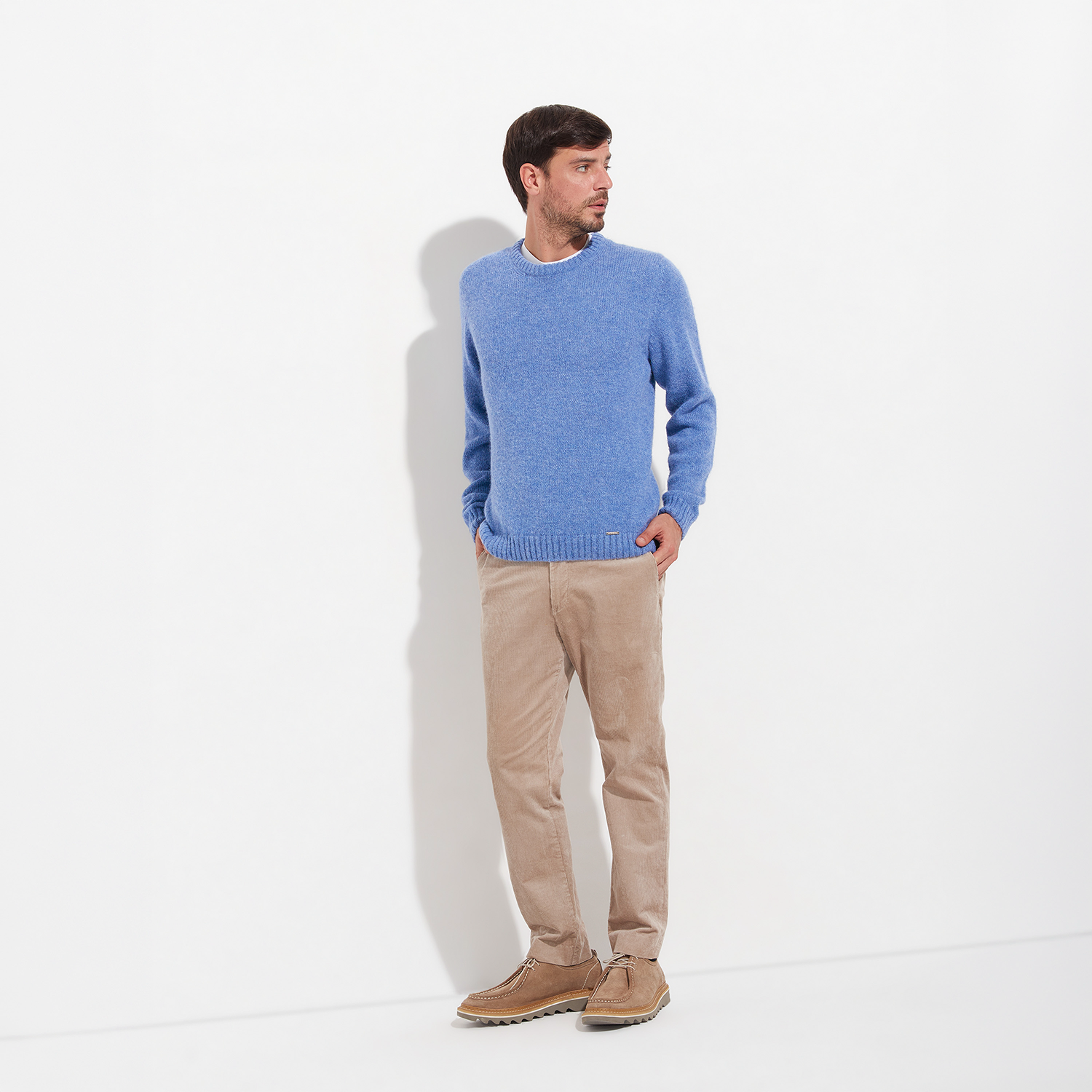 Man wearing a blue sweater and beige pants standing against a white background