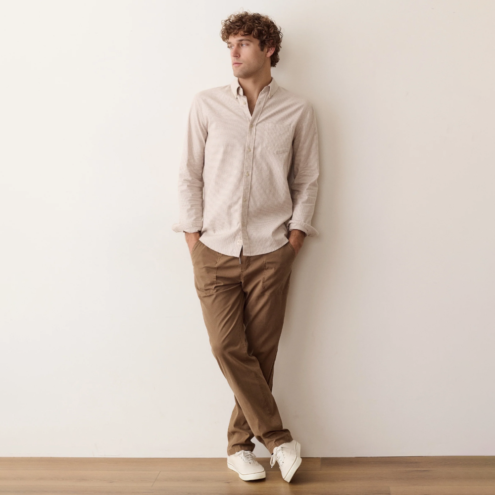 A man wearing a light beige long-sleeve button-down shirt, brown trousers, and white sneakers, leaning against a plain off-white wall on a wooden floor.