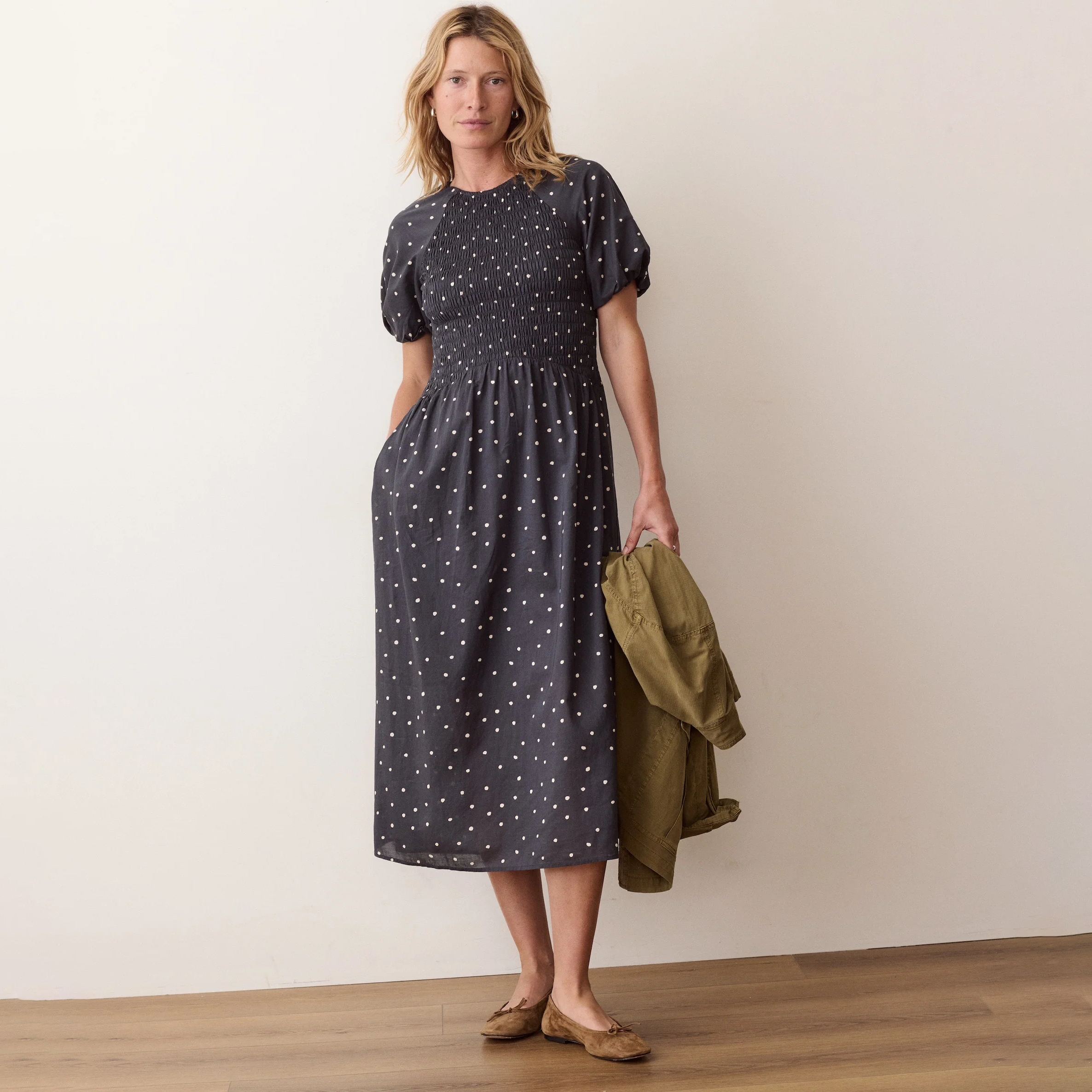 A woman stands in a studio wearing a dark navy midi dress with white polka dots, holding an olive green jacket. The background is a clean beige wall with a light wood floor.