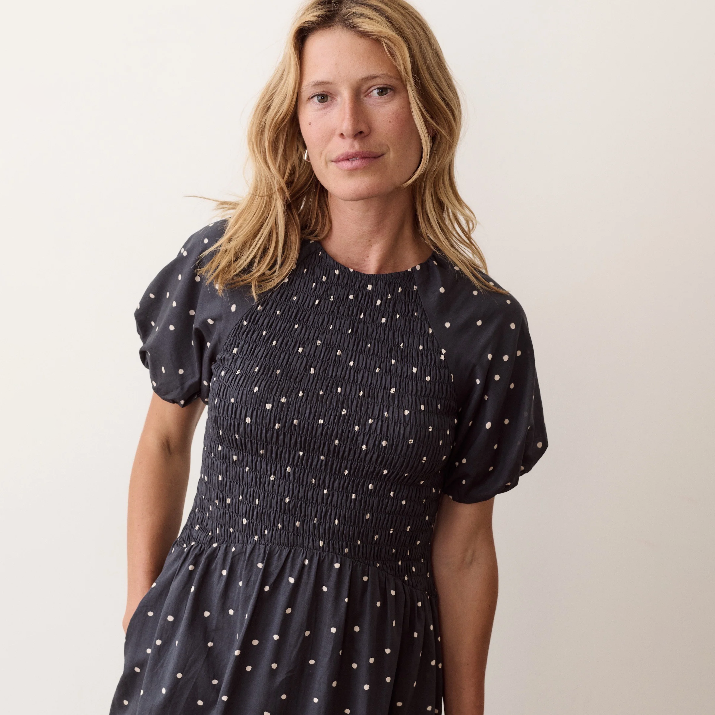 A woman wearing a navy smocked dress with white polka dots, photographed against a plain light beige studio background.
